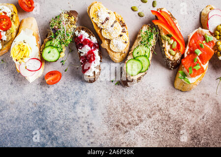 Toast mit verschiedenen Belägen auf grau-braunen Hintergrund geöffnet. Flach von Crostini mit Banane und Erdnussbutter, Pastete, Avocado, Lachs, Ei, Käse Stockfoto