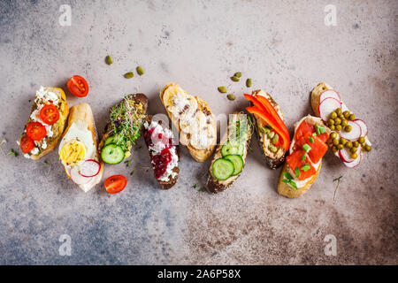 Toast mit verschiedenen Belägen auf grau-braunen Hintergrund geöffnet. Flach von Crostini mit Banane und Erdnussbutter, Pastete, Avocado, Lachs, Ei, Käse Stockfoto