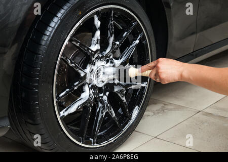 Ein männlicher Arbeitnehmer wäscht ein schwarzes Auto mit einer speziellen Bürste für Gegossene Räder und scheuert sich die Oberfläche zu glänzen in einem Fahrzeug mit Werkstatt. Auto Service in Stockfoto