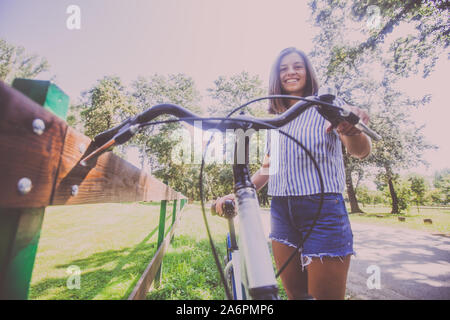 Portrait von Happy attraktive junge Frau in Jeans Shorts mit Fahrrad im Park im Sommer Tag. Low Angle View. Stockfoto