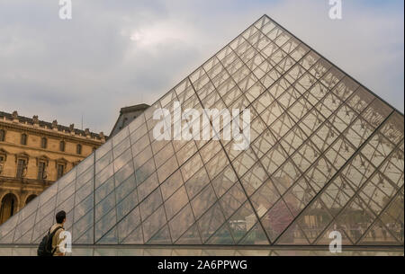 Ein männlicher Besucher mit einem Rucksack steht vor der Louvre Pyramide, sieht bis zu der Spitze und bewundert die zeitgenössische Glasbau in Paris. Stockfoto