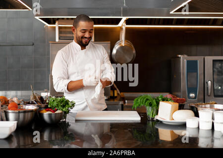 Junge attraktive männliche Cook bläst seine Schutzhandschuh Witze und hat Spaß in der professionellen Küche schöne schwarze Küchenchef in weiße Uniform gekleidet auf Stockfoto