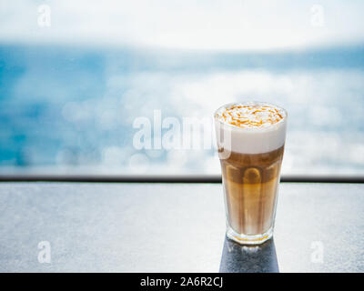 Hot Latte macchiato Kaffee mit Karamell auf der Tischplatte mit Blue Sea für den Hintergrund. Natürliche harte Tageslicht. Kopieren Sie Platz für Text oder Design Stockfoto