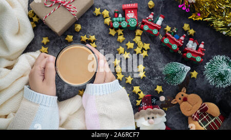 Hand mit heißen Getränk in der Wintersaison mit Weihnachten Hintergrund Dekorationen und Geschenkboxen auf Tisch Stockfoto