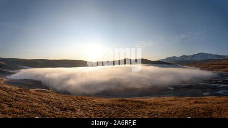 Erstaunlich herbst Panorama mit einem weißen flauschigen Decke von Nebel über einem großen See und flachen Seen, Gras und Bäume im Raureif, verschneite Berge und die Sonne Stockfoto