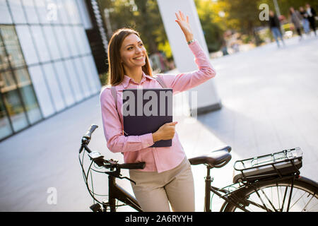 Glückliche junge Frau mit Dateien in den Händen stehen im Freien neben Elektrofahrrad Stockfoto