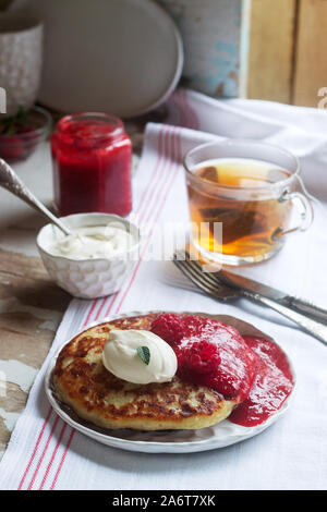 Quark-Pfannkuchen mit Erdbeersauce, saurer Sahne und Himbeeren und Kräutertee auf einem hellen Tisch. Rustikaler Stil. Stockfoto