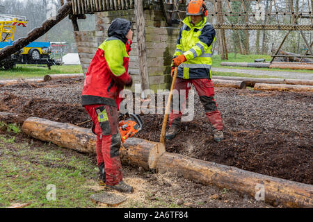 Junge Holzfäller arbeiten auf einem Spielplatz mit Baumhaus, schneiden Baumstamm mit Kettensägen in den regnerischen Tag. Stockfoto