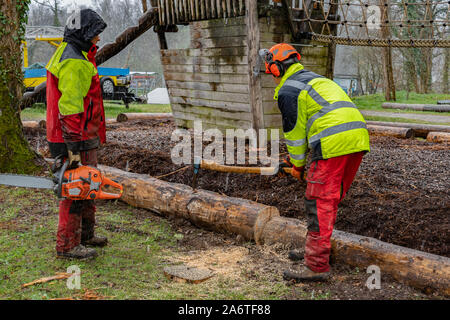 Junge Holzfäller arbeiten mit einer Axt auf einen Spielplatz, zweiten Logger wartet mit Kettensägen nach dem Schneiden Baumstamm im regnerischen Tag. Stockfoto