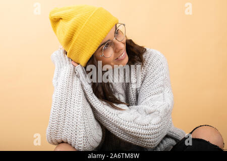 Eine junge Frau, gekleidet in einem Pullover, Gelb Hut und Brille sitzt auf dem Boden auf orangem Hintergrund und Träume Stockfoto