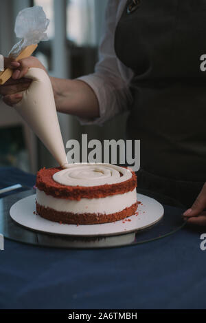 Pastry Chef drückt beige Creme auf Red Velvet Cake. Das Mädchen schmückt den Kuchen mit Sahne. Stockfoto