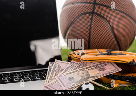 Nahaufnahme, Ansicht von Dollar Banknoten im Baseball Handschuh in der Nähe von Toy Fußball, Laptop und Basketball Ball isoliert auf weißem, Sport Konzept wetten Stockfoto