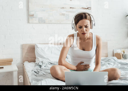 Betonte Frau in Kopfhörer Bauch berühren und mit Laptop auf dem Bett Stockfoto