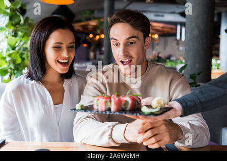 7/8-Ansicht von Kellner Halteplatte mit leckeren Sushi in der Nähe überrascht Paar in der Sushi Bar Stockfoto