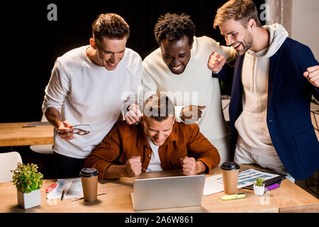 Fröhlicher junger Unternehmer in der Nähe von Laptop und zeigt Sieger Geste in der Nacht im Büro Stockfoto