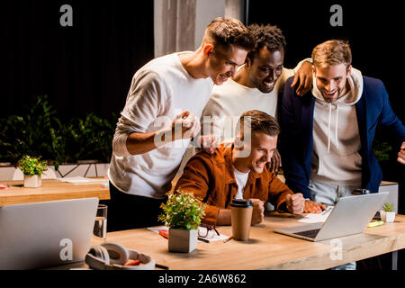 Glückliche junge Unternehmer auf der Suche nach Laptop und zeigt Sieger Geste in der Nacht im Büro Stockfoto