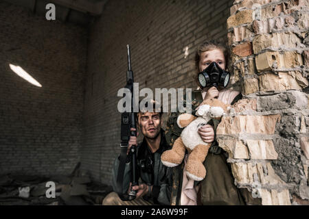 Kind in der Gasmaske holding Teddybär in der Nähe von stattlicher Mann mit Gewehr, post-apokalyptischen Konzept Stockfoto