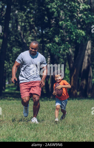 Fröhliche African American Boy mit rugby ball, während Spaß mit Vater im Park Stockfoto