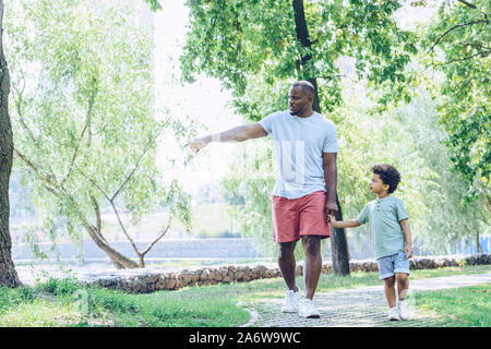 Junge afrikanische amerikanische Vater zeigt mit dem Finger während des Gehens mit entzückenden Sohn im Park Stockfoto