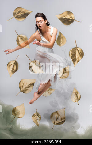 Schöne Ballerina in weißem Kleid tanzen durch fallende Blätter in der Nähe von grauer Rauch auf grauem Hintergrund umgeben Stockfoto