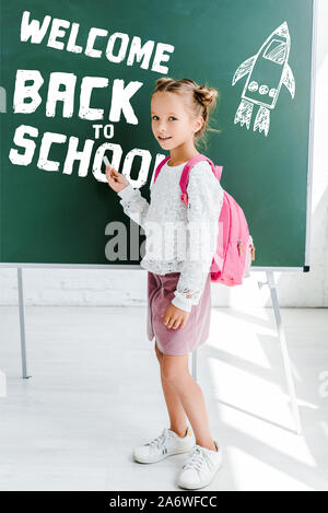 Süßes Schulmädchen mit Rucksack und mit Kreide in der Nähe willkommen zurück zu Schule Schriftzug auf der grünen Tafel Stockfoto