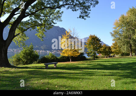 Wunderschöner Panoramablick auf das grüne Lecco Seeufer mit großen Bäumen und Holzbänken zur Entspannung und auf das Wasser des Garlate Sees an herbstlichen sonnigen Tagen. Stockfoto