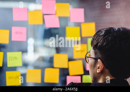 Asiatische Geschäftsmann auf der Suche und Schreiben auf Kurznotiz für Brainstorming Ideen auf klare Whiteboard in moderne Büro. Analyse und Synthese Stockfoto