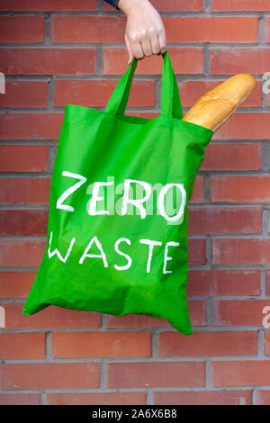 Grünen eco Beutel mit weißem Text null Abfall in der Hand auf einem Hintergrund von Red brick wall. Lange französisches Brot Sticks aus der Tasche. Vertikale. Stockfoto