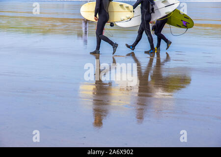 Surfer, die Surfbretter tragen, an einem nassen Strand in Polzeath, Cornwall, Großbritannien, spazieren gehen Stockfoto