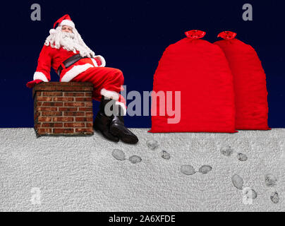 Santa Claus sitzt auf dem Schornstein an einem verschneiten Dach mit großen Taschen voller Weihnachtsgeschenke, Hintergrund von dunklem Blau mit Sternen. Stockfoto