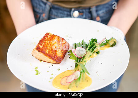 Der Kellner hält in seinen Händen Lachs Filet und Steak mit jungen Brokkoli garniert. Restaurant Menü, eine Reihe von Fotos von verschiedenen Gerichten Stockfoto