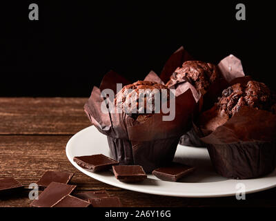Stillleben mit braunen Muffins mit Kakao in einer Verpackung von Backpapier, Stücke Schokolade auf einem weißen Porzellanteller und alten hölzernen Tisch. Schwarz Stockfoto