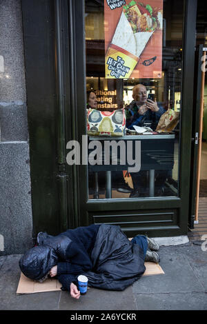 Rauer Schlaf liegt auf der Straße außerhalb einer Fast-Food-Kette, während die Menschen im Inneren der warmen Speisen am Fenster, Central London, Großbritannien sitzen Stockfoto