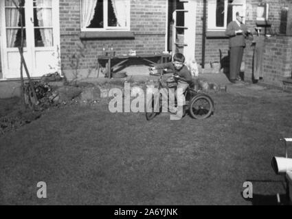 1950er Jahre, historisch, ein kleiner Junge, der auf seinem Dreirad auf dem Gras in einem Garten eines Hauses reitet, England, Großbritannien. Stockfoto