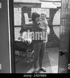 1960er Jahre, historisch, eine junge Mutter, die ihr Kind draußen in einem Hinterhof hält, neben ihr, einem traditionellen Kutschenwagen, in England, Großbritannien. Stockfoto