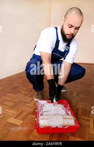 Bauarbeiter tun beenden Renovierung im Apartment. Maler mit Handschuhen hält weiße Walze Pinsel in der hand und Malen im Container- und roten Schüssel Stockfoto