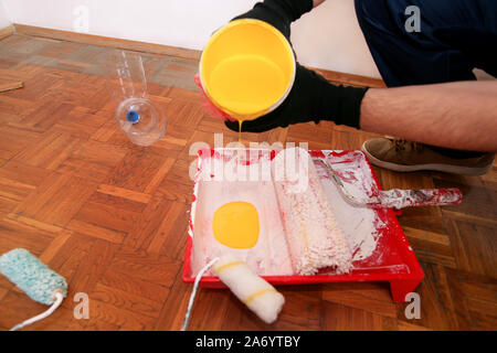 Bauarbeiter tun beenden Renovierung im Apartment. Maler mit schwarzen Handschuhen gießt gelbe Farbe von der Schaufel in die roten Schüssel für Farbe mischen. Stockfoto