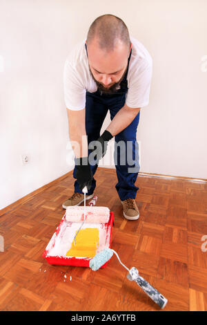 Bauarbeiter und Maler mit Handschuhen hält Bürstenrolle Farbe in der Hand und Mixe gelbe Farbe im Container- und roten Schüssel für Farbmischung. Stockfoto