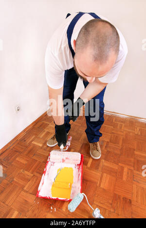 Bauarbeiter und Maler mit Handschuhen hält Bürstenrolle Farbe in der Hand und Mixe gelbe Farbe im Container- und roten Schüssel für Farbmischung. Stockfoto