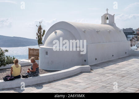 Eine winzige kleine weiße Wäsche Kirche Kapelle mit Kuppel und Glockenturm mit Kreuz Kruzifix und zwei Touristen auf kurze Wand Oia Santorini sitzen Stockfoto