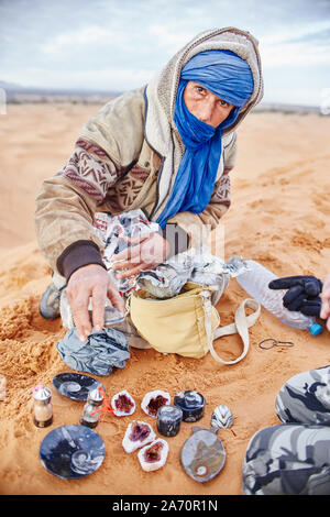 Berber Mann in der Wüste Sahara bietet seine eigenen Souvenirs und Edelsteine. Bedouin sitzen auf dem Sand auf der Düne. Marokko, Merzouga, 16. Dezember Stockfoto