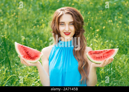 Porträt der schönen jungen Frau trägt blaues Kleid halten zwei Scheiben Wassermelone auf Gras Hintergrund. Stockfoto