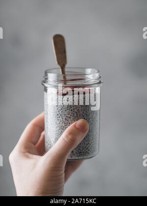 Chia Pudding in der Frau die Hand auf grauem Hintergrund. Weibliche Hand halten Glas Glas mit Chia Samen puding Goji Beeren serviert. Kopieren Sie Platz für Text. Vertikale. Stockfoto