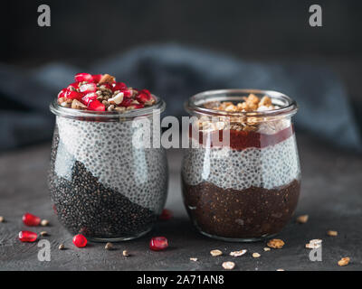 Zwei chia Pudding in Gläsern auf dunklen Tisch. Zwei Farben anthrazit Chia puding mit Granatapfel-, Mandel-, Hanf samen. Schokolade chia Pudding mit Marmelade und Müsli. Kopieren Sie Platz. Gesunde Übernachtung Frühstück Stockfoto