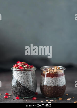 Zwei chia Pudding in Gläsern auf dunklen Tisch. Zwei Farben anthrazit Chia puding mit Granatapfel-, Mandel-, Hanf samen. Schokolade chia Pudding mit Marmelade und Müsli. Kopieren Sie Platz. Gesunde Übernachtung Frühstück Stockfoto