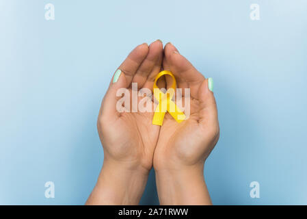 Gelb Gold Ribbon in den Händen eines Mädchens auf einem blauen Hintergrund. Kinder Krebs Konzept. Sstsein Monat. Krebs im Kindesalter Tag Februar, 15. Stockfoto