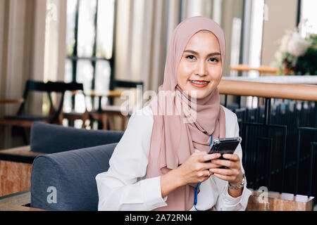 Optimistisch asiatische Frau im hijab lächelnd und Suchen an der Kamera, während im Cafe sitzen und Surfen social media auf dem Mobiltelefon Stockfoto