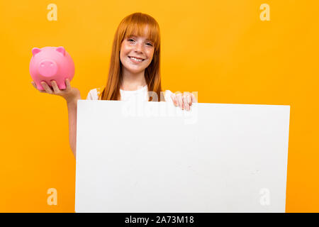 Rothaarige nettes Mädchen mit einem breiten Grinsen auf orangem Hintergrund mit einem Sparschwein in Händen, leeres Formular für Text, Banken und Versicherungen Konzept Stockfoto