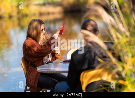Junge Frau mit einem selfie auf ihr Mobiltelefon als sitzt sie Entspannung in ein Restaurant im Freien mit Blick auf einen ruhigen See mit ihrem Freund Stockfoto