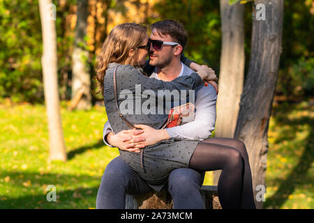 Gerne schwangere junge Frau sitzt auf dem Schoß ihres Mannes, wie Sie, im Freien in einem Herbst Park entspannen Stockfoto